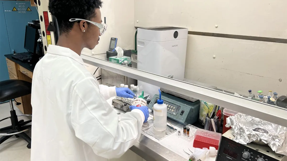 A man wearing a lab coat, safety glasses and blue gloves stands at a laboratory fume hood, running a research experiment. The workspace contains various lab items, including bottles, water samples and electronic instruments.