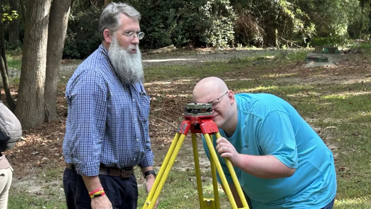 Two men stand outdoors on a grassy area beneath trees, using a yellow tripod mounted surveying instrument. One man is looking through the device while the other stands nearby observing.