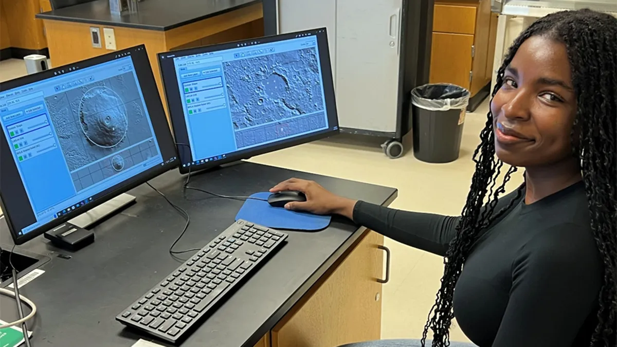 A woman sits at a laboratory workstation using a computer with two monitors that display detailed photographs of the surface of planet Mars.