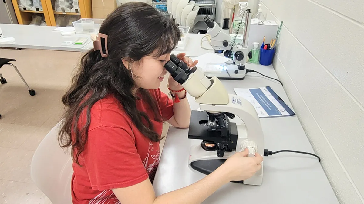 A woman sits at a laboratory workstation and looks into a compound microscope. One hand adjusts the focus knob while the microscope is illuminated from below.