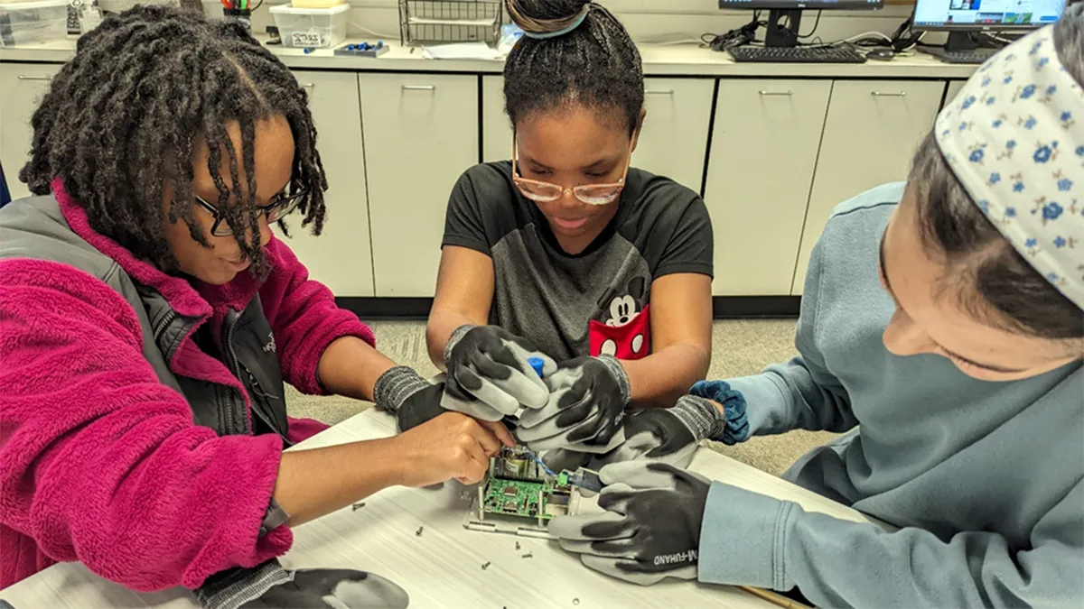 Three students sit at a table working together to assemble a seismograph. All three wear protective gloves and use small tools to assemble the components. Loose screws, tools and device parts are scattered on the table.