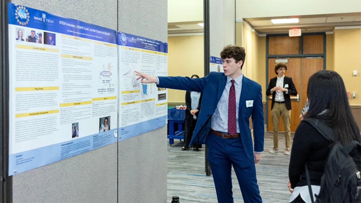 A man wearing a suit stands beside his research poster and points to a section of it while speaking to another person, who is facing the poster. The poster contains text, charts, graphics and small photos describing his research. The scene takes place in a conference-style room.