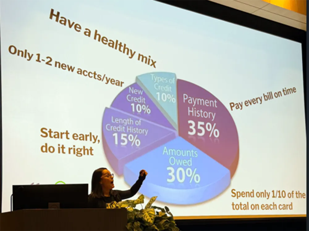 A money management expert speaks on stage with a pie chart graphic displayed behind her.