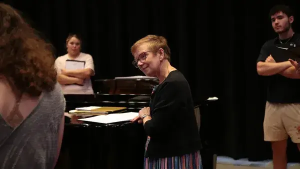 Associate Professor Katharine Boyes stands next to a piano, with a couple members of the Wake Tech chorus in the background.