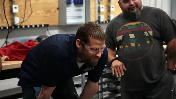 Instructor Stephen Tracey, wearing clear plastic safety goggles, bends over to examine work being done by students during a Building Automation Technology class as another student looks on.