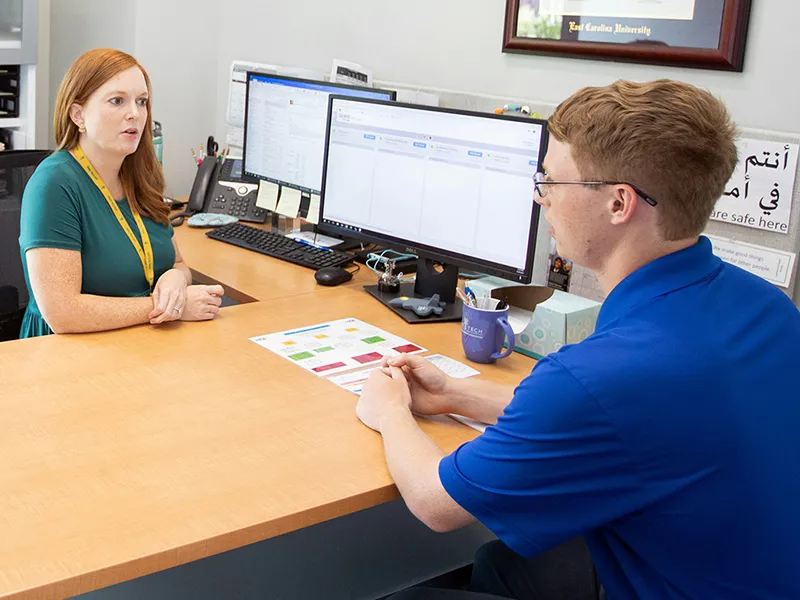 image of a student in a blue shirt speaking to a woman sitting at a desk