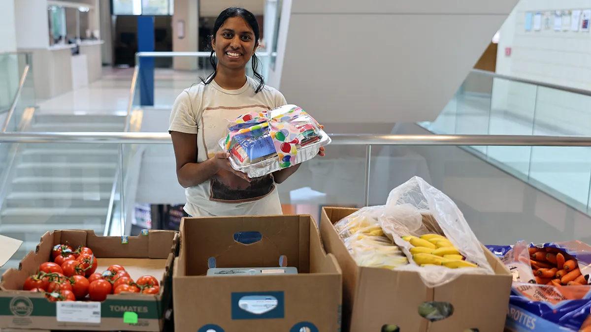 A woman poses behind boxes of tomatoes, eggs, bananas and carrots.