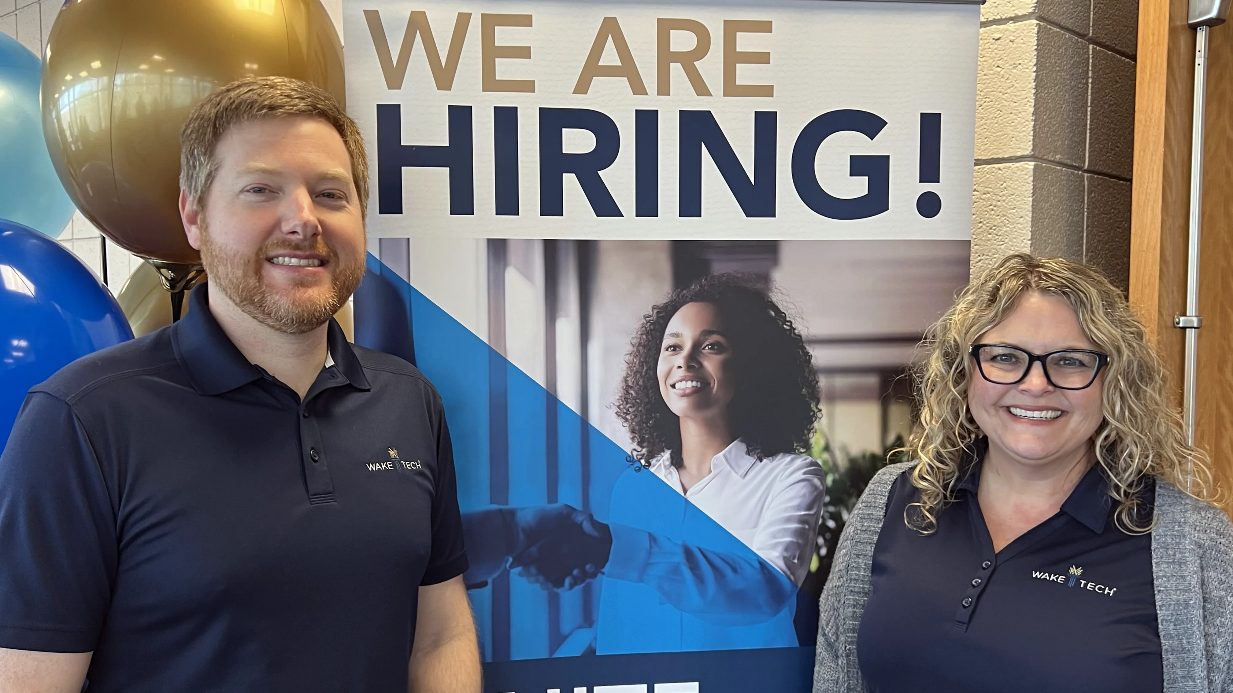 A man and a woman stand in front of a poster that says, &quot;We Are Hiring!&quot;