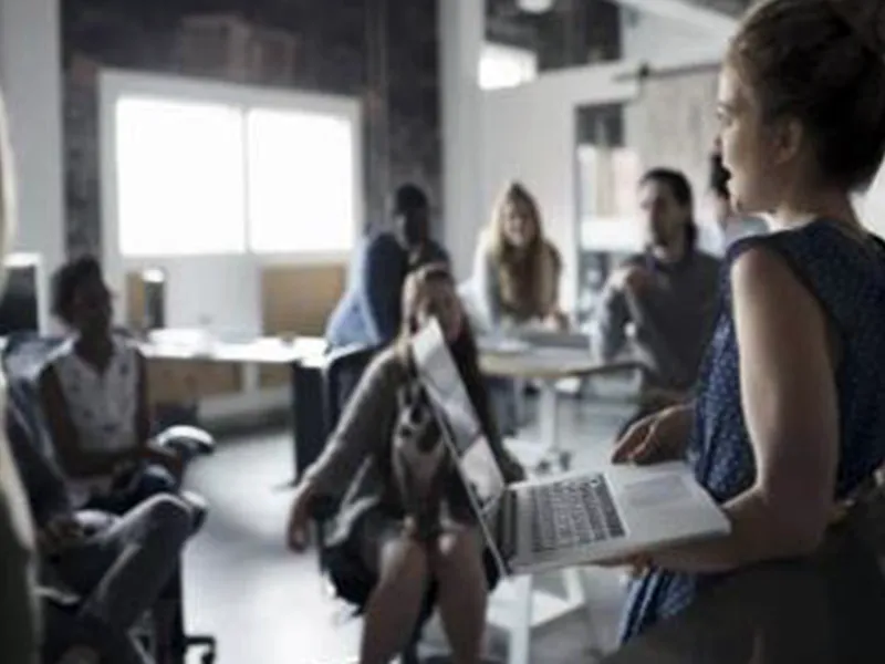 A woman holding a laptop speaks to a group in a classroom.