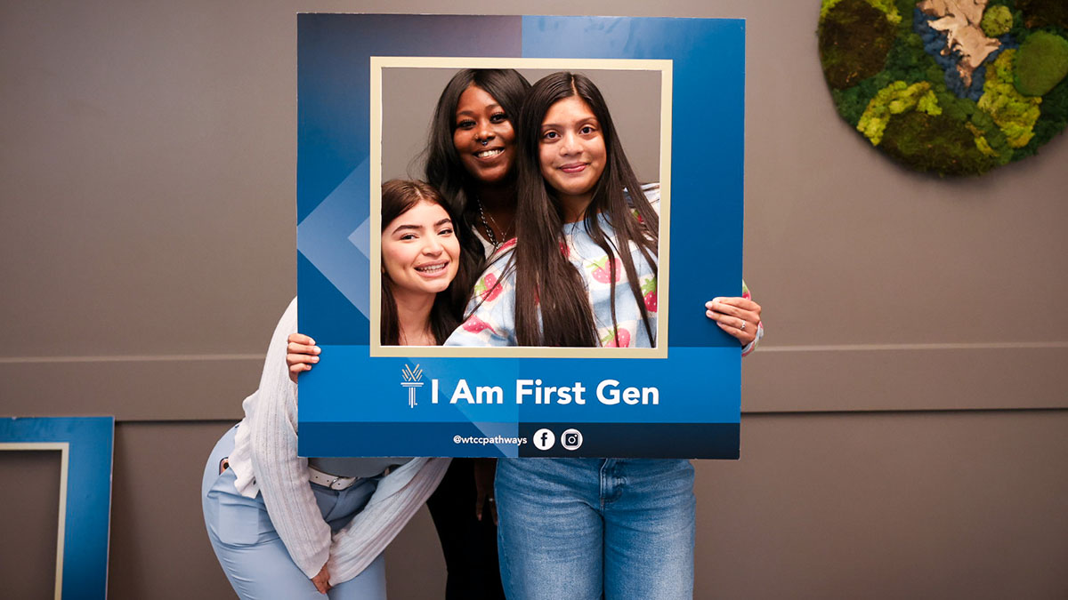 Three female Wake Tech students pose for a photo behind an enlarged blue frame with the words "I am first-gen" on it.