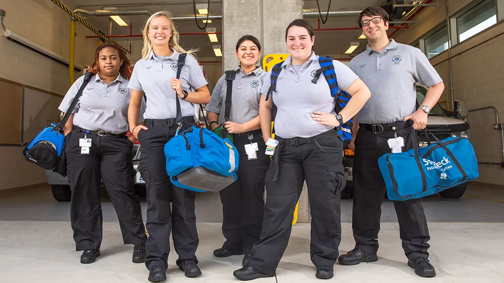 Five EMT students pose for a photo in an ambulance bay.