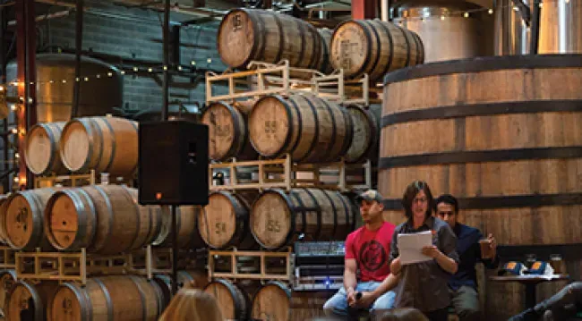 Three men sit in a warehouse next to stacks of wooden beer barrels.