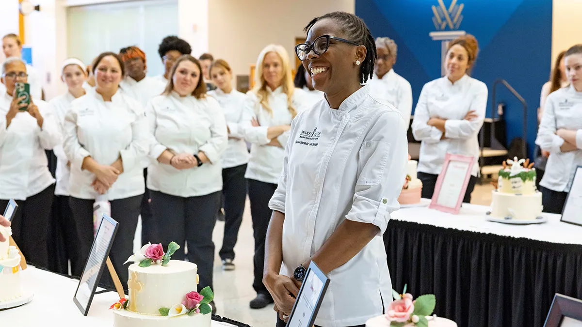 A smiling female student dressed in a white chef's coat stands beside a decorated tiered cake on a table. Other students dressed in white chef coats are in the background.