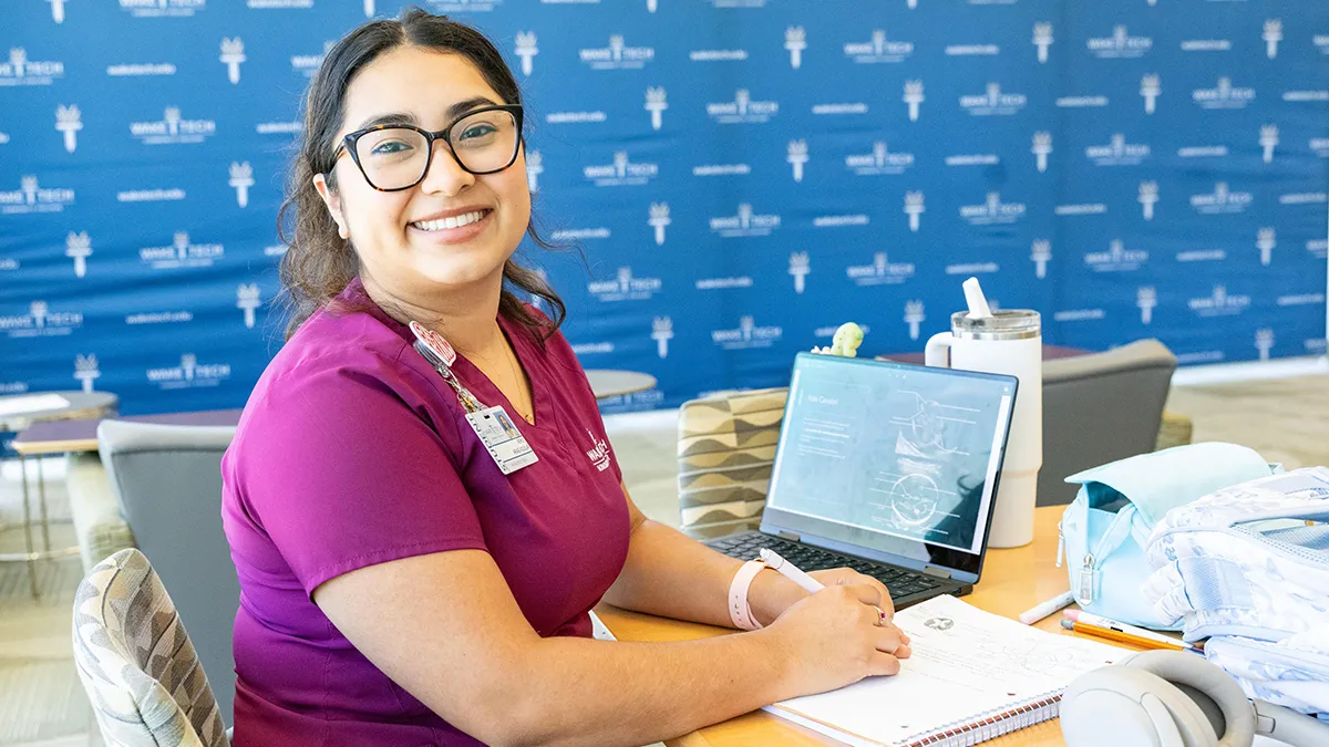 A smiling student sitting at a desk with an open notebook and laptop computer