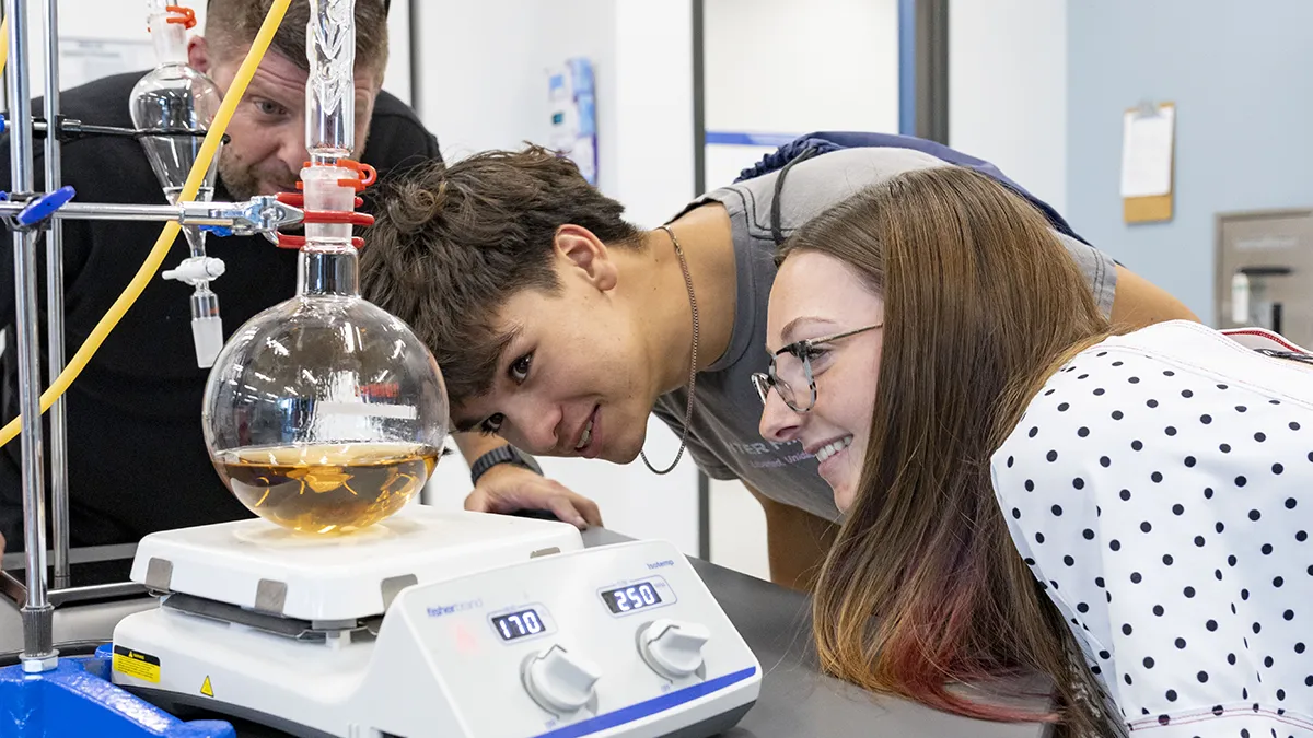 Two men and a woman watch as an amber-colored liquid is heated and spun in a flask in a chemistry lab.