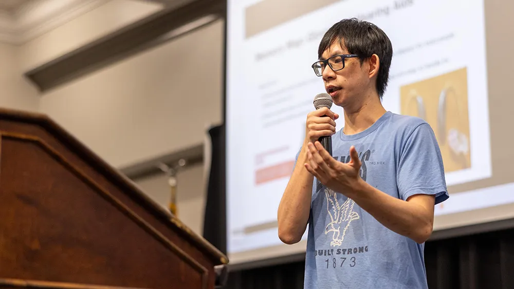 A male student speaks into a microphone with a slide projected on a screen behind him.