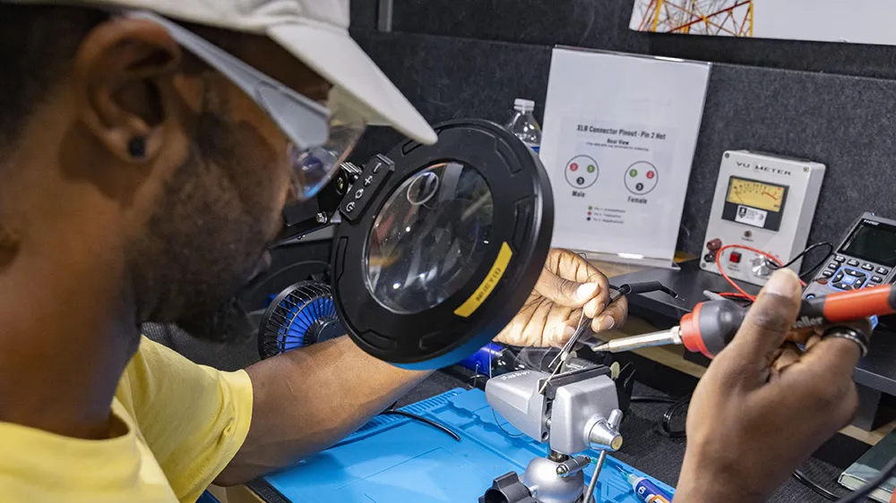 A student uses a large magnifying glass to see the electrical wires he's working with at a work station.