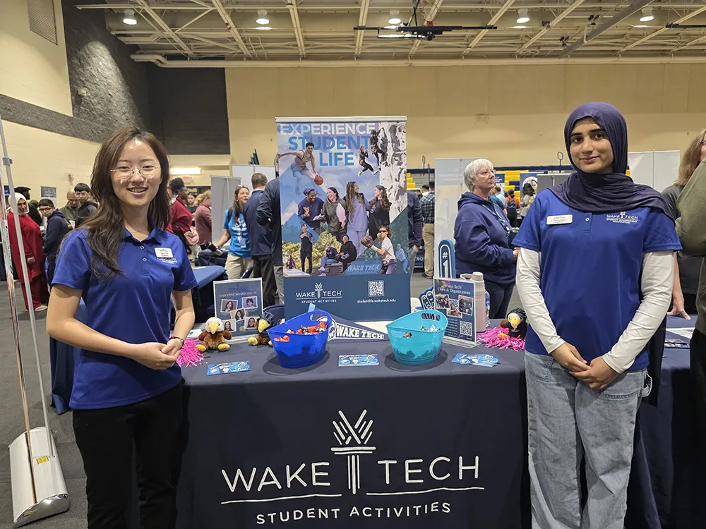 Two Wake Tech students staff a Student Activities table at an open house, greeting visitors at an academic and resource fair inside a campus building.
