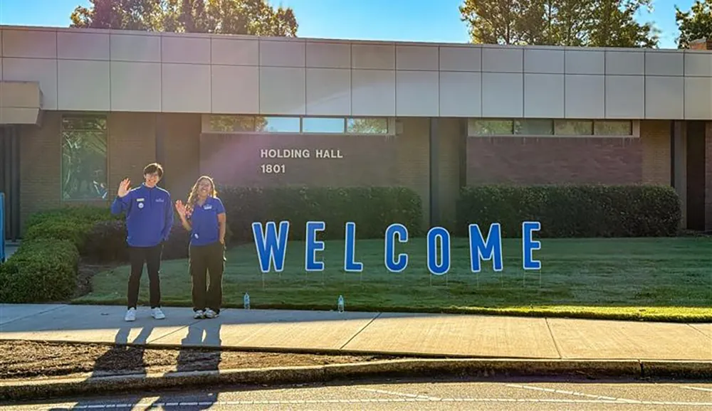 Two students stand waving next to a sign that says "welcome" outside a Wake Tech building.