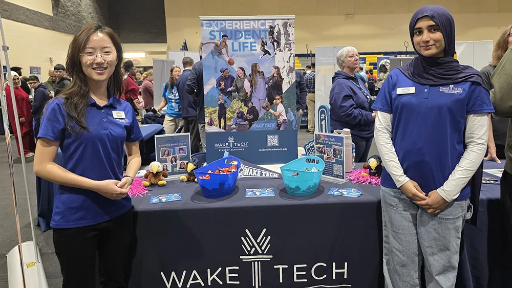 Two Wake Tech students staff a Student Activities table at an open house, greeting visitors at an academic and resource fair inside a campus building.
