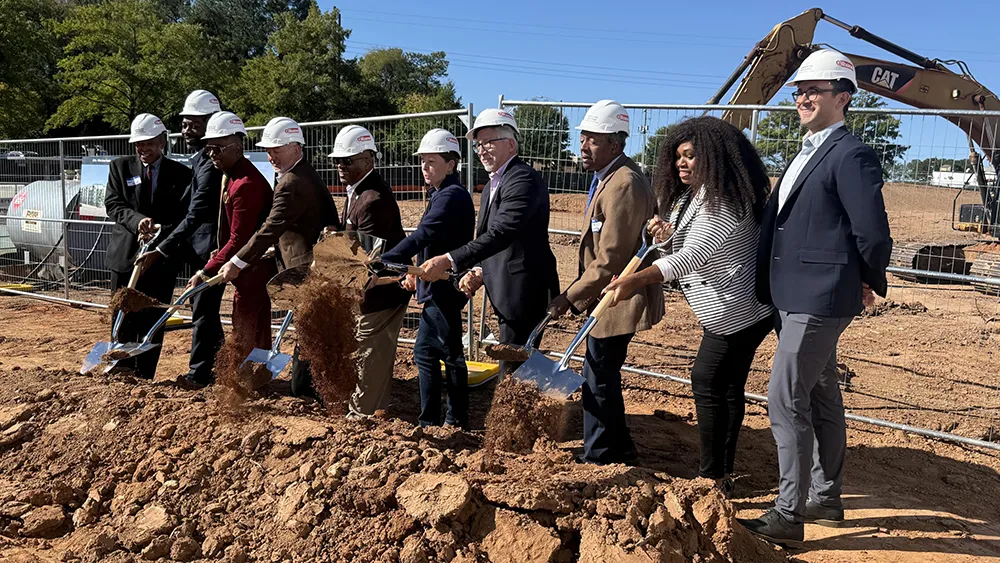Ten people in white hardhats use shovels to pitch dirt during a groundbreaking ceremony for a planned affordable housing community.