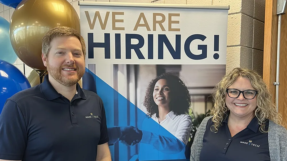 Wake Tech staff stand in front of a "We're Hiring" banner