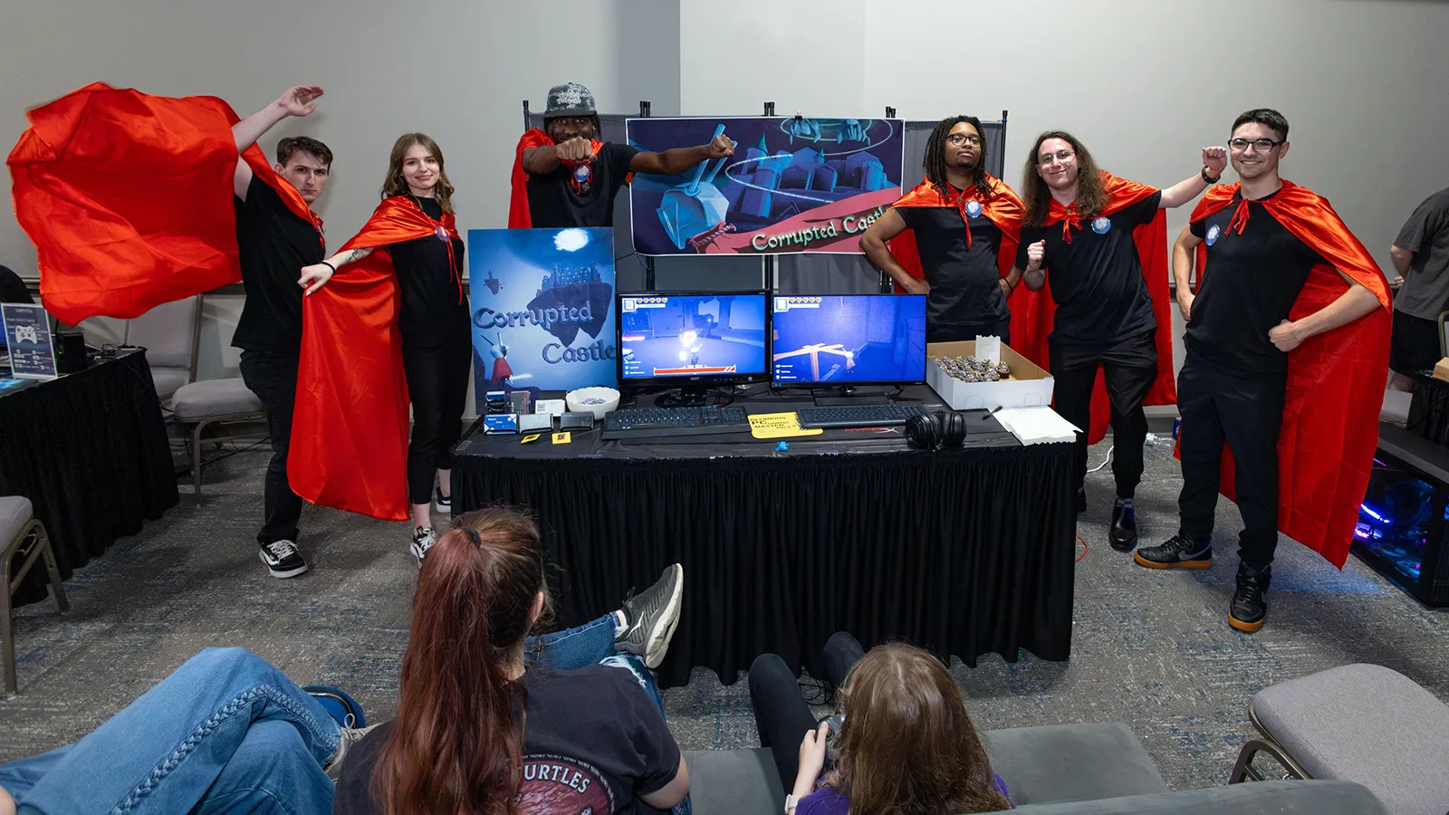 Six Wake Tech students dressed in black with red capes stand behind a table with a sign and computer screens displaying their "Corrupted Castle" video game.