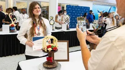 A male student with a gold medal around his neck displays a certificate as he and two American Culinary Federation judges pose for a photo.