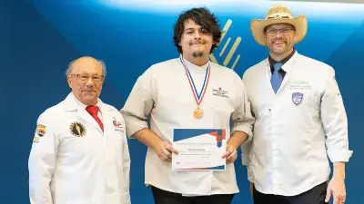 A male student with a gold medal around his neck displays a certificate as he and two American Culinary Federation judges pose for a photo.