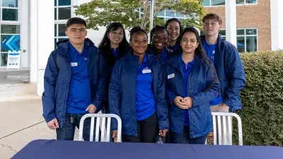 A group seven male and female students in matching blue shirts and windbreakers pose for a photo.