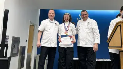 Two men in white chef coats stand next to woman with a medal around his neck holding a certificate.