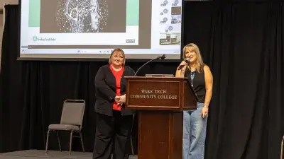 Two women stand behind a podium with a slide projected on a screen behind them.