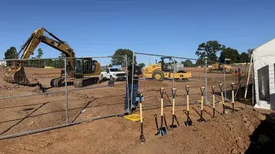 A chain-link fence separates construction equipment in the background from a row of shovels in the foreground on land that will become an affordable housing development.