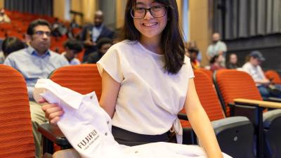 Female student seated in an auditorium holding a white lab coat with a Fujifilm logo before Wake Tech’s Biotechnology White Coat Ceremony, with other attendees visible in the background.