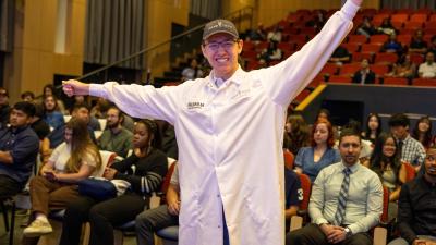 Female student seated in an auditorium holding a white lab coat with a Fujifilm logo before Wake Tech’s Biotechnology White Coat Ceremony, with other attendees visible in the background.