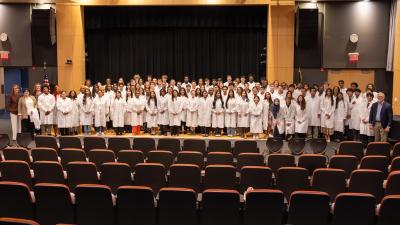 Female student seated in an auditorium holding a white lab coat with a Fujifilm logo before Wake Tech’s Biotechnology White Coat Ceremony, with other attendees visible in the background.