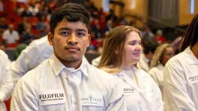 A seated female student holds a white lab coat with a Fujifilm logo on her lap.
