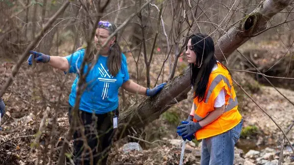 A woman in a blue T-shirt and a young woman in an orange vest stand in a heavily wooded area. The woman in the T-shirt is pointing to something.
